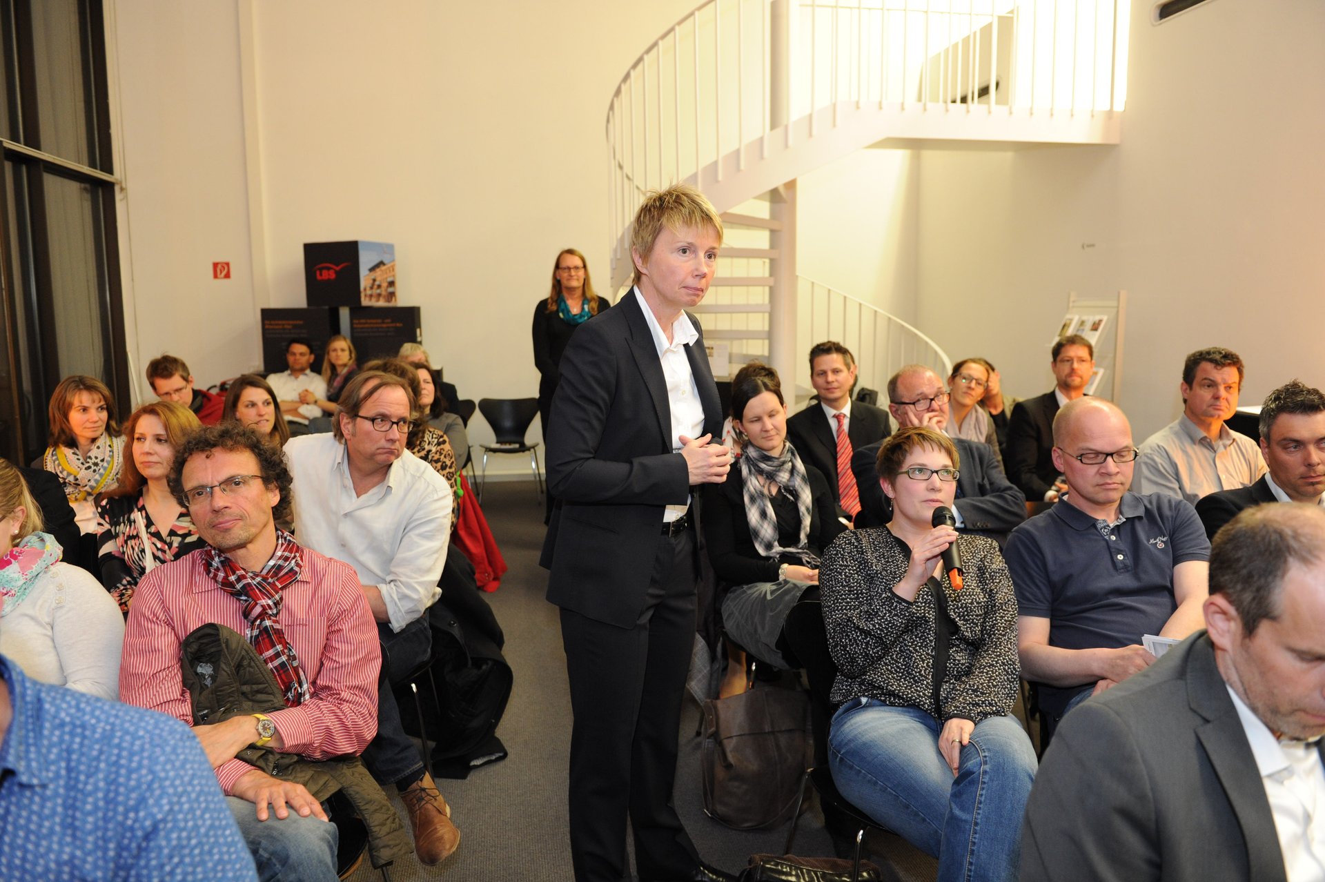 Bild der sitzenden Besucher sowie der Moderatorin Alexandra May. Ein Frau aus dem Publikum spricht in das Mikrofon in ihrer Hand. (Foto: Kristina Schäfer)