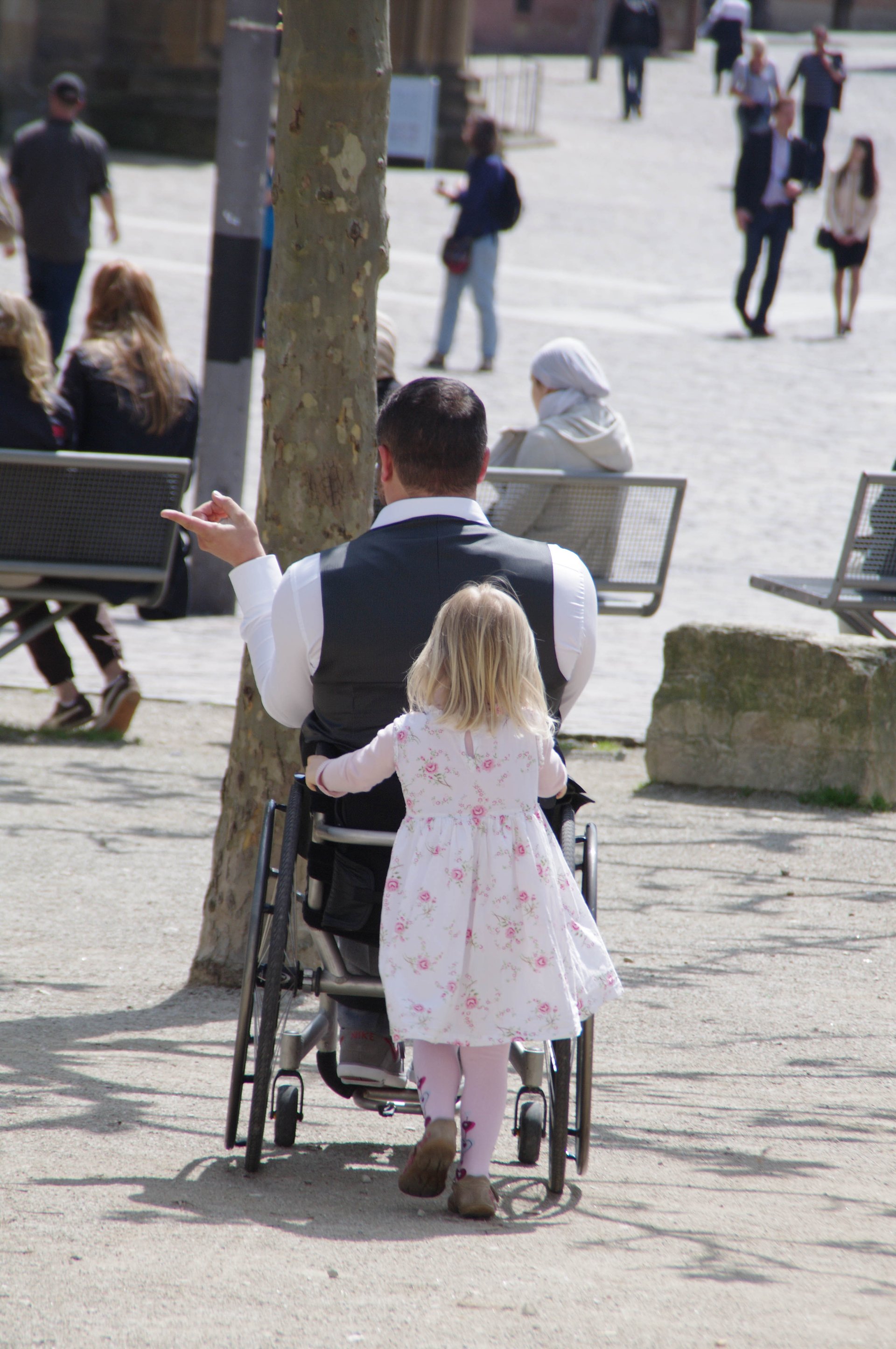 Ein kleines Mädchen in einem geblümten Kleid schiebt einen Mann im Rollstuhl auf einem öffentlichen Platz in Richtung eines Baumes. Der Mann zeigt mit einem Finger nach Links, um die Richtung anzugeben, in die das Mädchen ihn schieben soll. (Foto: Architektos)