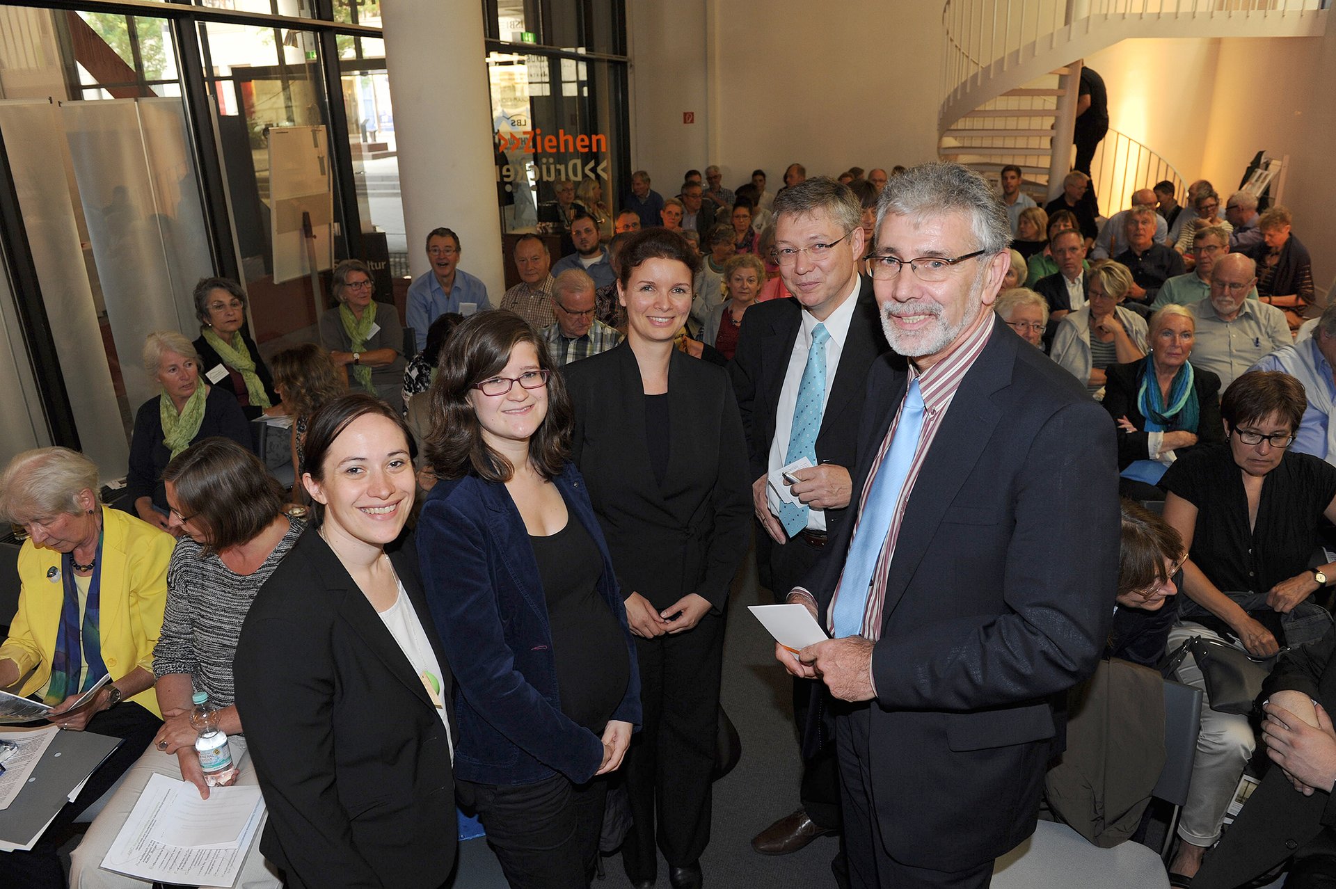 Personengruppe mit fünf Personen, aufgestellt im Halbkreis, im Hintergrund weitere, sitzende Personen. (Foto: Kristina Schäfer)