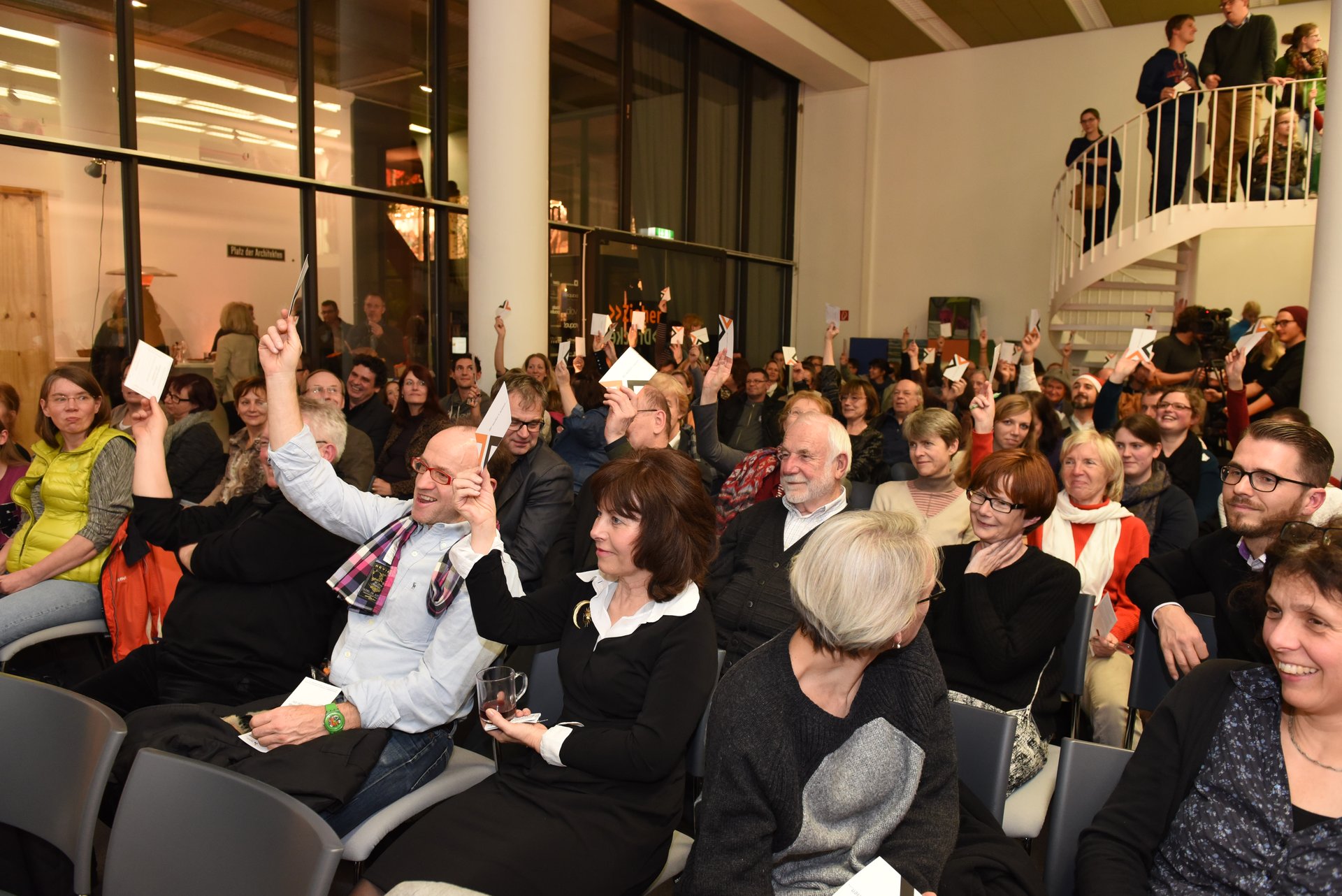 Zahlreiche sitzende Besucher halten jeweils eine Karte mit ausgestrecktem Arm zur Abstimmung in die Höhe. Zahlreiche sitzende Besucher halten jeweils eine Karte mit ausgestrecktem Arm zur Abstimmung in die Höhe.