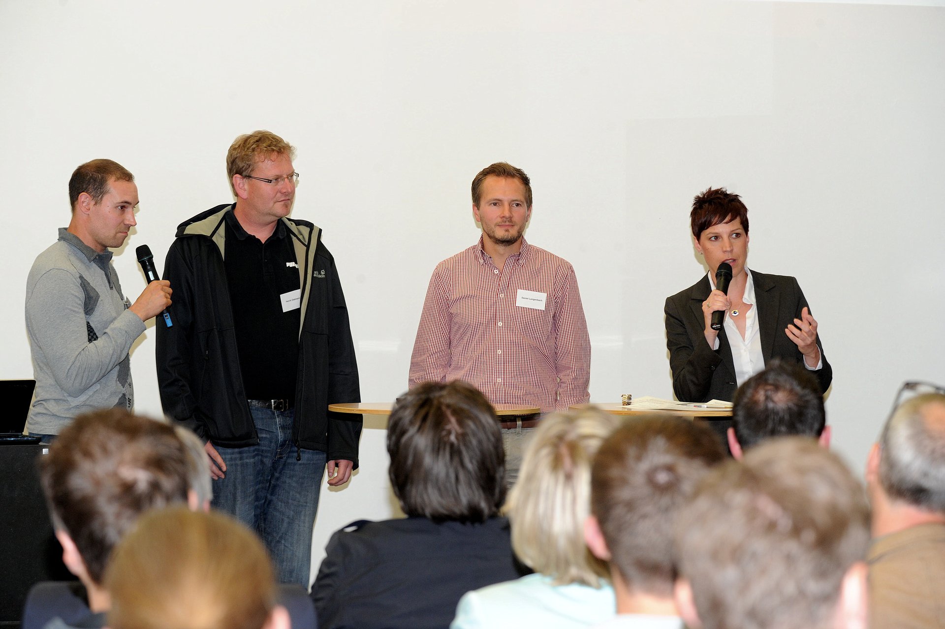 Drei Männer und eine Frau, nebeneinander stehend, teilweise mit Mikrofon in der Hand, im Vordergrund die Hinterköpfe des Publikums (Foto: Kristina Schäfer)