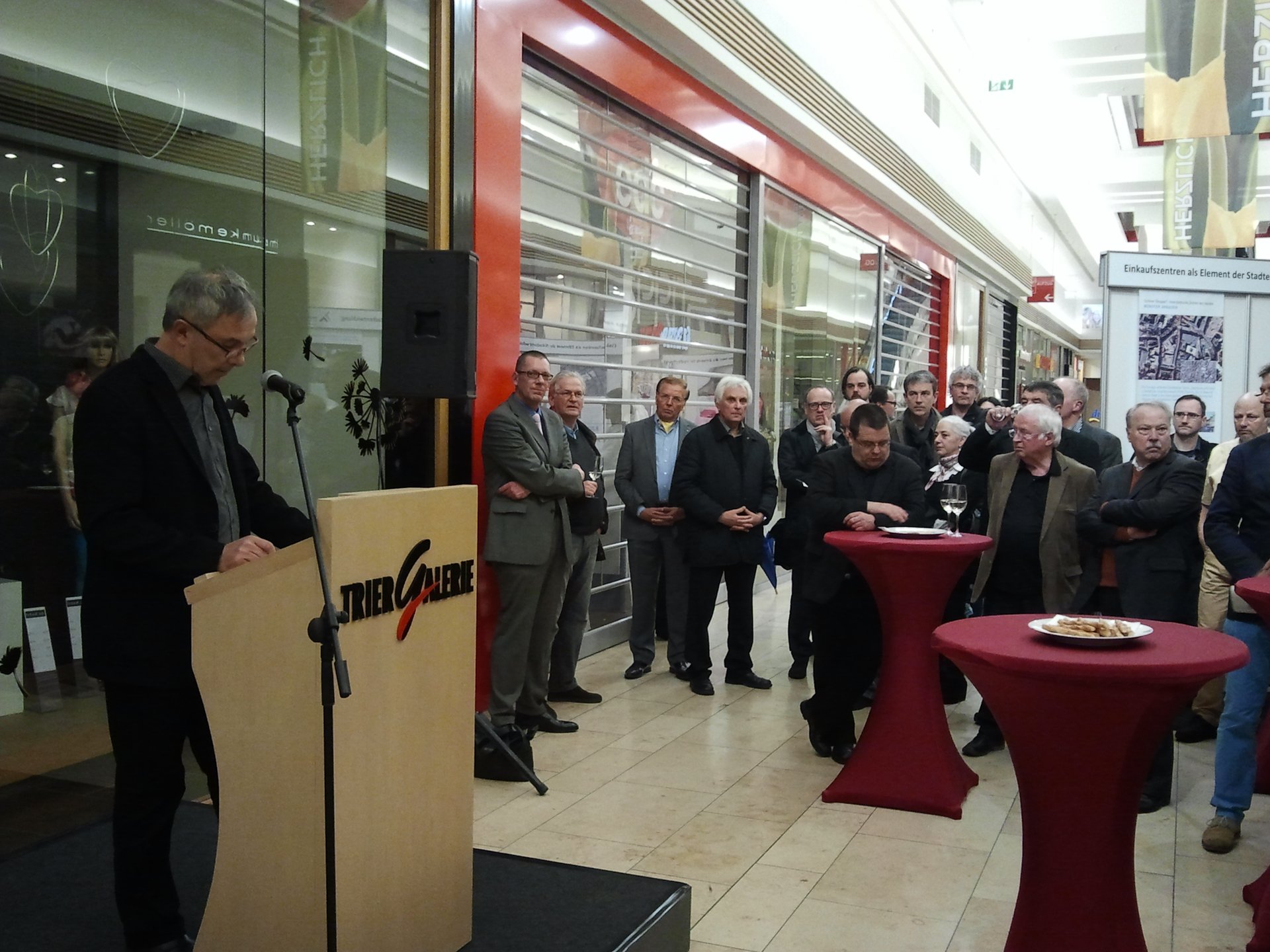 Innenaufnahme der TRIER GALERIE, Kammervizepräsident Manfred Müller rechts am Stehpult, in der Mitte und rechts im Bild zahlreiche Zuhörer, teilweise an Stehtischen stehend. (Foto: Birgit Brüggen)
